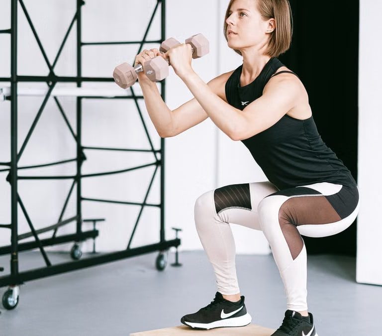 A woman in sportswear performs a squat with dumbbells indoors, embodying strength and healthy living.